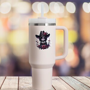May include: A white travel mug with a black handle and a sticker of a cartoon girl wearing a black cowboy hat and a black dress with a floral design. The girl has dark hair and dark eyes. The sticker is surrounded by a floral design with red and purple flowers.