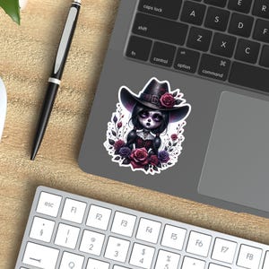 May include: A sticker featuring a dark-haired girl with a sugar skull face wearing a black cowboy hat with red flowers. She is surrounded by purple and red roses and vines.