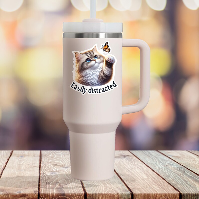 May include: A white travel mug with a handle, featuring a cartoon cat looking up at a butterfly with the text "Easily distracted".