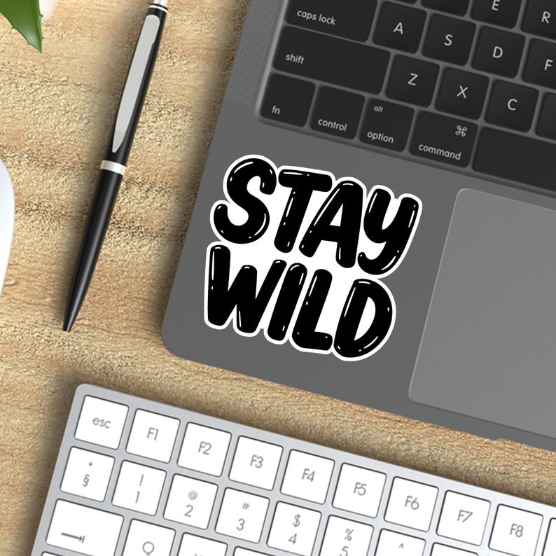 May include: A black and white sticker with the text "STAY WILD" in a bold, handwritten font. The sticker is placed on a laptop.