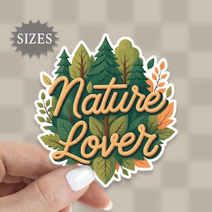 May include: A sticker with the words "Nature Lover" in a stylized, cursive font. The design features a forest scene with green trees, leaves, and orange accents. The sticker is held by a hand, and the word "SIZES" is in a gray starburst shape.
