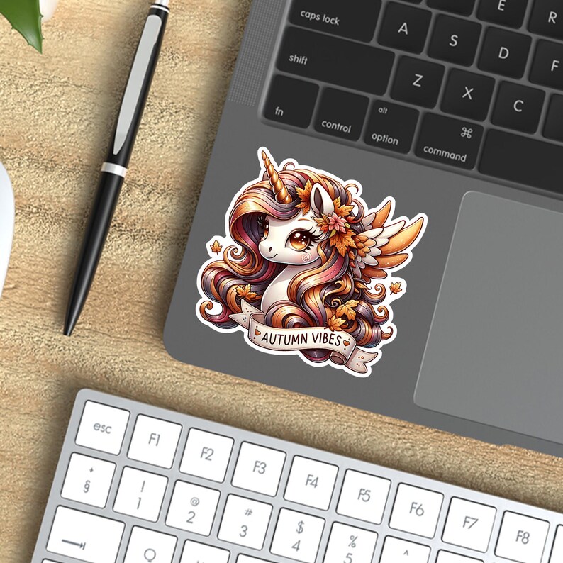 K&ouml;nnte beinhalten: Ein Aufkleber mit einem Cartoon-Einhorn mit einer M&auml;hne in Herbstfarben, verziert mit Herbstbl&auml;ttern und -bl&uuml;ten. Der Aufkleber tr&auml;gt den Text "AUTUMN VIBES" auf einem Banner. Der Aufkleber befindet sich auf einem Laptop.