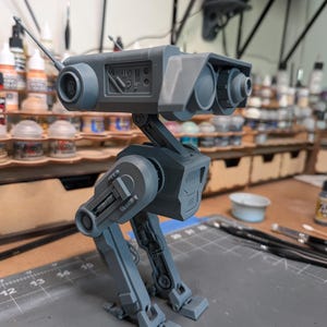 Könnte beinhalten: Ein graues, zweibeiniges Robotermodell mit detailliertem Design. Das Modell hat einen kastenförmigen Kopf mit zwei kreisförmigen, augenähnlichen Merkmalen und einer Antenne. Die Beine sind beweglich, und das Modell steht auf einer grauen Oberfläche. Das Modell ist wahrscheinlich ein Sammlerstück oder ein Kunstwerk.