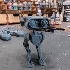 Könnte beinhalten: Ein graues, 3D-gedrucktes Modell eines Roboters mit einem kastenförmigen Kopf, zwei großen augenähnlichen Merkmalen und einer Antenne. Der Roboter hat einen rechteckigen Körper und zwei Beine. Das Modell steht auf einer grauen Oberfläche mit Farbtöpfen und Pinseln im Hintergrund.