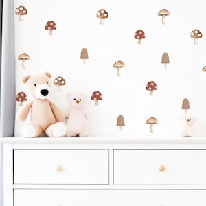 May include: A collection of brown and tan mushroom wall decals arranged on a white wall above a white chest of drawers. The set includes various mushroom shapes and sizes, featuring spotted caps and plain caps, alongside two plush teddy bears.