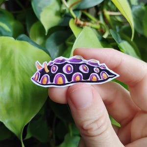 May include: A die-cut sticker featuring a stylized illustration of a sea slug. The creature has a black body with a repeating pattern of purple and orange circular spots, a white outline, and two small orange antennae protruding from the front.