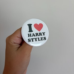 May include: A circular white pin-back button featuring a black letter I, a red heart symbol, and the black text HARRY STYLES printed in a bold, sans-serif font. The design is centered on the glossy surface of the round pin badge.