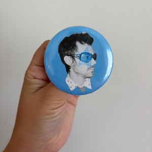 May include: A circular pinback button featuring a blue background and a black and white portrait of a person wearing blue-tinted sunglasses and a collared shirt with a small star graphic overlaid on the face and glasses area.