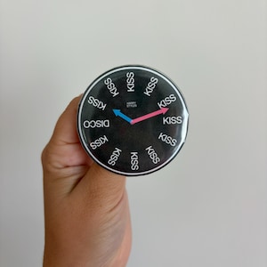 May include: A circular pin-back button featuring a black background with the word KISS repeated in white around the perimeter, the word DISCO, and the text HARRY STYLES in the center. It has a blue hour hand and a pink minute hand like a clock.