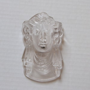 May include: A clear glass sculpture of a woman's head with detailed hair and a draped garment. The sculpture is a decorative piece.