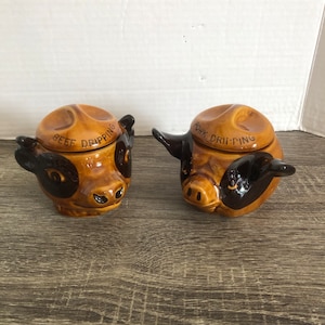 May include: Two ceramic containers shaped like a cow and a pig. The containers are brown and black with white accents. The cow container has the text "Beef Dripping" on the lid. The pig container has the text "Pork Dripping" on the lid.