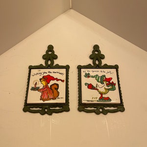May include: Two green metal pot holders with a white ceramic tile insert. The tile on the left has a cartoon squirrel wearing a red and white striped pajamas and a red hat holding a candle and a plate of holly. The tile on the right has a cartoon duck wearing a green and white striped hat and red boots holding a gift. The tile on the left says "Wishing you the merriest!" and the tile on the right says "Tis the season to be jolly!"