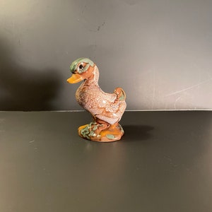 May include: A ceramic duck figurine. The duck has a brown body with white speckles, a green head, and a yellow beak. The base is brown with green and yellow accents. The figurine is on a brown surface.