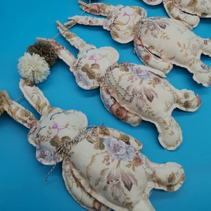 May include: A garland of four beige fabric bunnies with floral patterns, each with a brown and white pom-pom on top. The bunnies are strung together with twine and have wooden clothes pegs.
