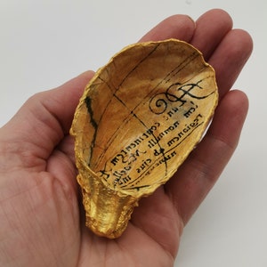 May include: A gold-painted seashell with a black inscription that reads "Legions of the night, will you come?" The shell is held in a hand.