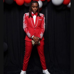 May include: A red suit with white accents and embellishments. The suit includes a blazer, pants, and a bow tie. White sneakers complete the outfit. Red, black, and silver balloons are in the background.