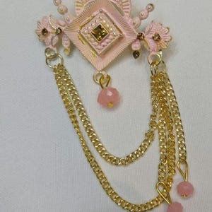 以下が含まれることがあります： 幾何学模様と垂れ下がったチェーンが付いたピンクとゴールドの装飾的なブローチ。ブローチの中央には、ピンクとゴールドの正方形があり、その中にさらに小さなゴールドの正方形があります。ブローチはピンクとゴールドの花で囲まれており、ピンクのビーズがぶら下がったゴールドのチェーンが3本付いています。