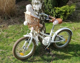 Puede incluir: Una bicicleta blanca con una cesta de mimbre en el manillar. Un animal de peluche está sentado en la cesta. La bicicleta está aparcada junto a un abedul.