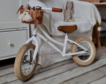 Puede incluir: Bicicleta de equilibrio blanca con una cesta de mimbre marrón y un sillín de cuero marrón. La bicicleta está en un suelo de madera clara en una habitación con un sofá blanco y una ventana.