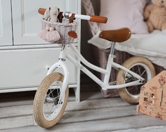 Puede incluir: Bicicleta de equilibrio blanca con manillares marrones y una cesta de mimbre con un pompón rosa. La bicicleta tiene un asiento de cuero marrón y neumáticos beige. La bicicleta está en una habitación con un armario blanco y una silla de mimbre con un cojín rosa.