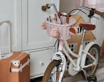 Puede incluir: Una bicicleta blanca con una cesta de mimbre rosa y un asa de cuero marrón. La cesta tiene un juguete de conejo rosa dentro y pompones rosas en los lados. La bicicleta está aparcada junto a una maleta marrón.
