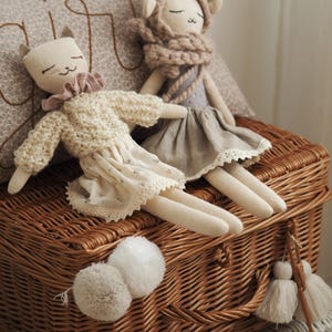 Peut inclure: Deux poupées en tissu faites à la main avec des visages brodés. L'une porte un pull en tricot torsadé crème et une jupe à fleurs avec bordure en dentelle. L'autre porte une robe grise avec un ourlet en dentelle et une capuche en tricot épais.