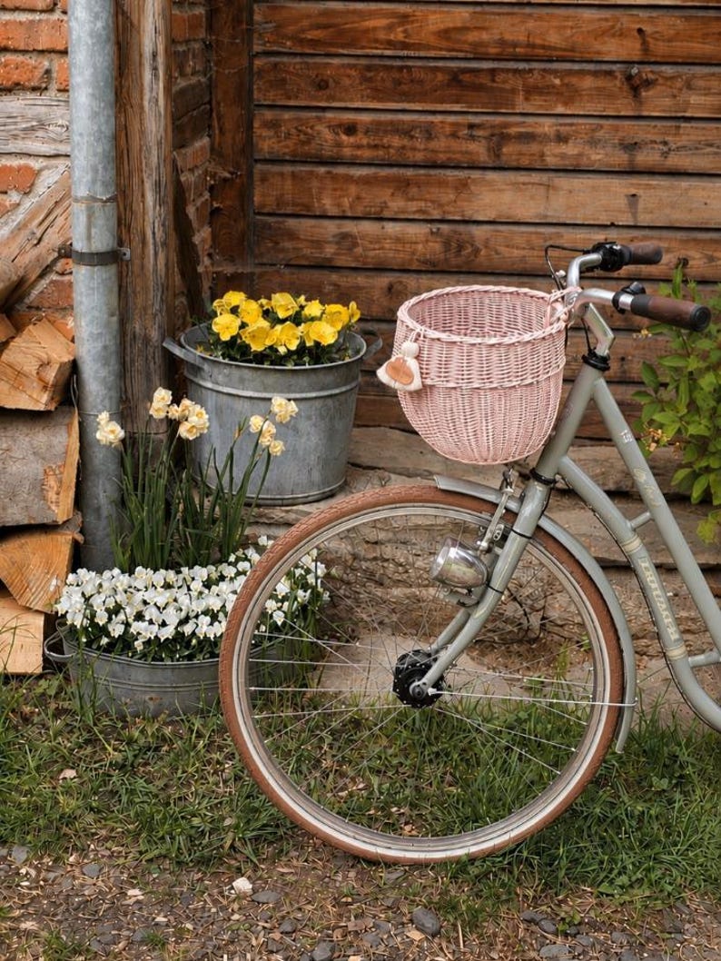 Puede incluir: Una bicicleta gris claro con una cesta redonda de mimbre trenzado rosa p&aacute;lido sujeta al manillar. La cesta cuenta con un peque&ntilde;o adorno de borla beige. La bicicleta tiene pu&ntilde;os de goma marr&oacute;n y un faro de metal plateado en el cuadro.