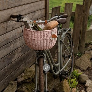 Puede incluir: Una cesta de bicicleta de mimbre trenzado de color rosa claro montada en el manillar de una bicicleta gris. La cesta contiene una hogaza de pan, un manojo de esp&aacute;rragos verdes y un objeto de papel estampado. Una etiqueta de cuero cuelga.