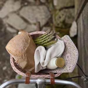 Puede incluir: Una cesta de bicicleta tejida de color rosa claro con correas de cuero marr&oacute;n que contiene una hogaza de pan en una bolsa de papel marr&oacute;n, un manojo de esp&aacute;rragos verdes, un tejido de punto color crema doblado, una botella de vidrio con tapa dorada y un cuaderno floral.