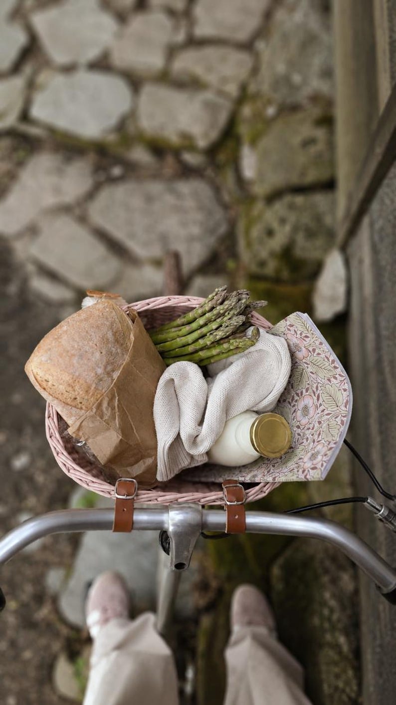 Puede incluir: Una cesta de bicicleta tejida de color rosa claro con correas de cuero marr&oacute;n que contiene una hogaza de pan en una bolsa de papel marr&oacute;n, un manojo de esp&aacute;rragos verdes, un tejido de punto color crema doblado, una botella de vidrio con tapa dorada y un cuaderno floral.