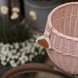 Puede incluir: Una cesta de bicicleta de mimbre tejido en color rosa claro con una peque&ntilde;a etiqueta de cuero marr&oacute;n en forma de nube con el texto Little Dreams grabado, decorada con una borla de hilo color crema atada al borde con un cordel fino.