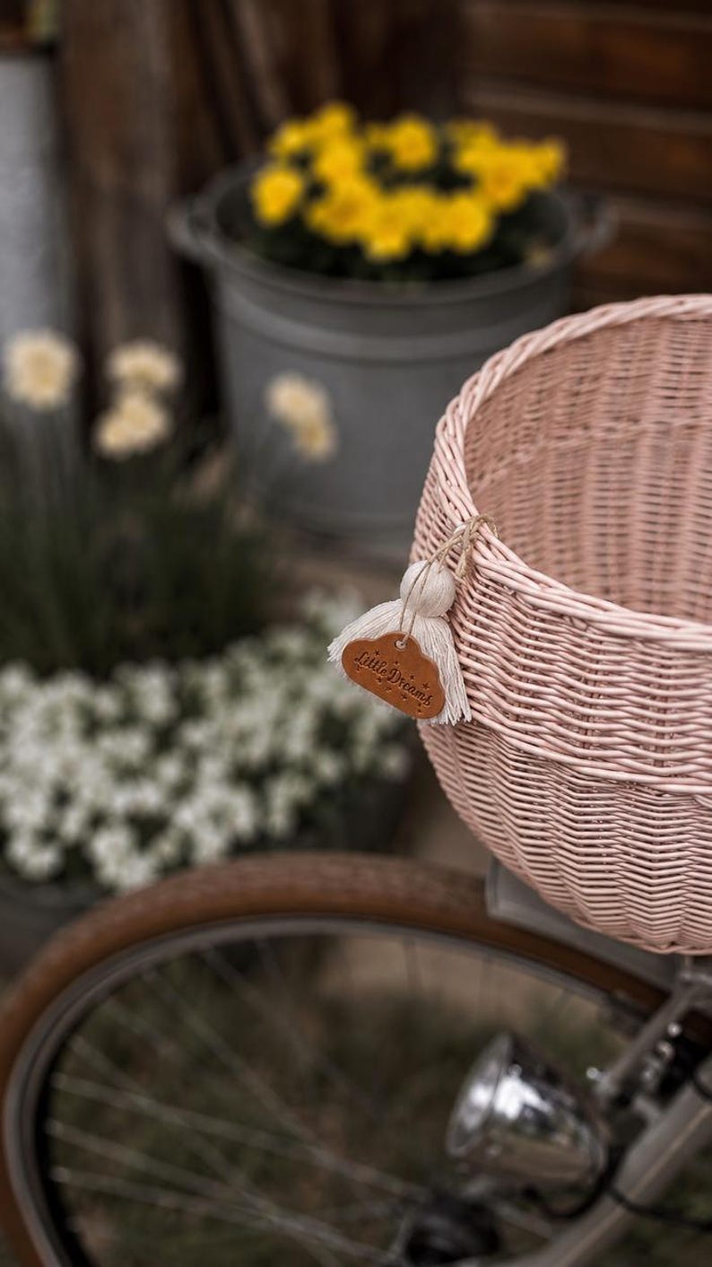 Puede incluir: Una cesta de bicicleta de mimbre tejido en color rosa claro con una peque&ntilde;a etiqueta de cuero marr&oacute;n en forma de nube con el texto Little Dreams grabado, decorada con una borla de hilo color crema atada al borde con un cordel fino.