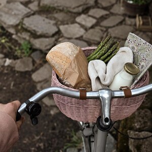 Puede incluir: Una cesta de bicicleta tejida de color rosa claro sujeta a un manillar plateado, que contiene una hogaza de pan en una bolsa de papel marr&oacute;n, un manojo de esp&aacute;rragos verdes, un pa&ntilde;o color crema doblado, una botella de leche y un cuaderno floral.