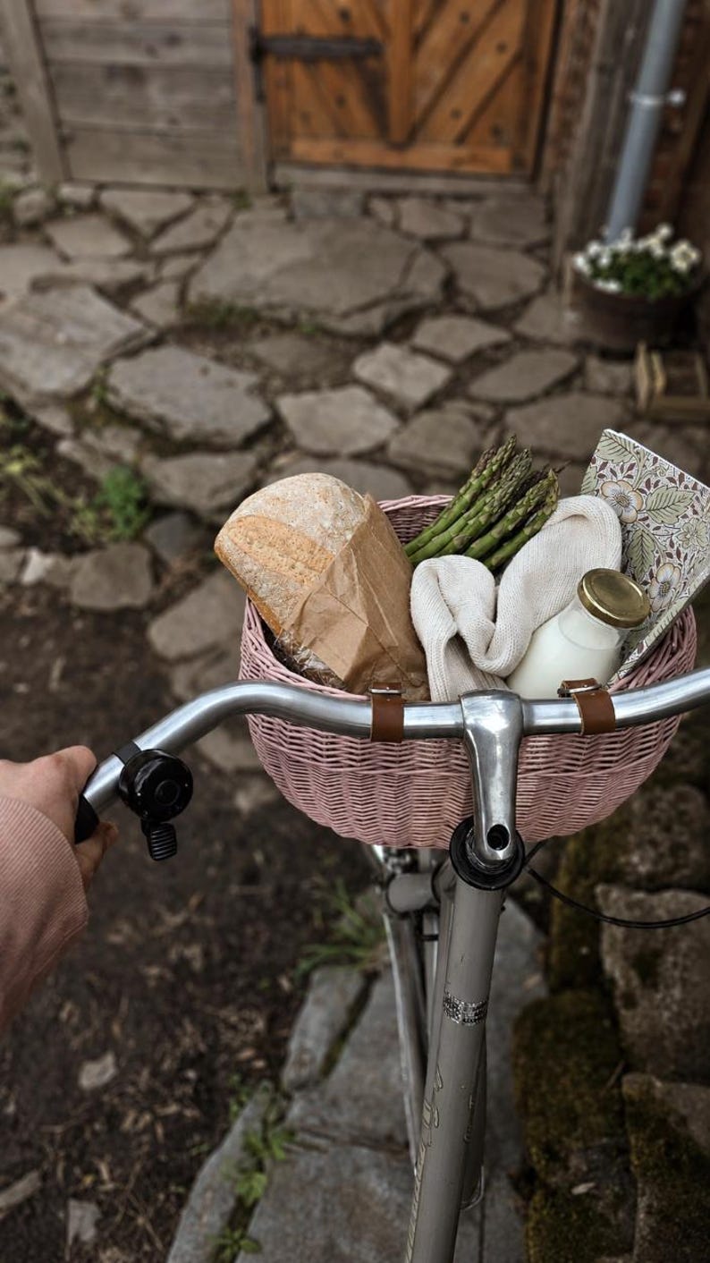 Puede incluir: Una cesta de bicicleta tejida de color rosa claro sujeta a un manillar plateado, que contiene una hogaza de pan en una bolsa de papel marr&oacute;n, un manojo de esp&aacute;rragos verdes, un pa&ntilde;o color crema doblado, una botella de leche y un cuaderno floral.
