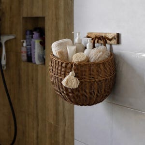 Könnte beinhalten: Ein runder, brauner geflochtener Weidenkorb, der an einer hölzernen Wandhalterung hängt. Der Korb enthält weiße Handtücher, eine Holzbürste, eine beige Quaste und mehrere weiße Kunststoff-Pumpflaschen mit verschiedenen Pflegeprodukten.
