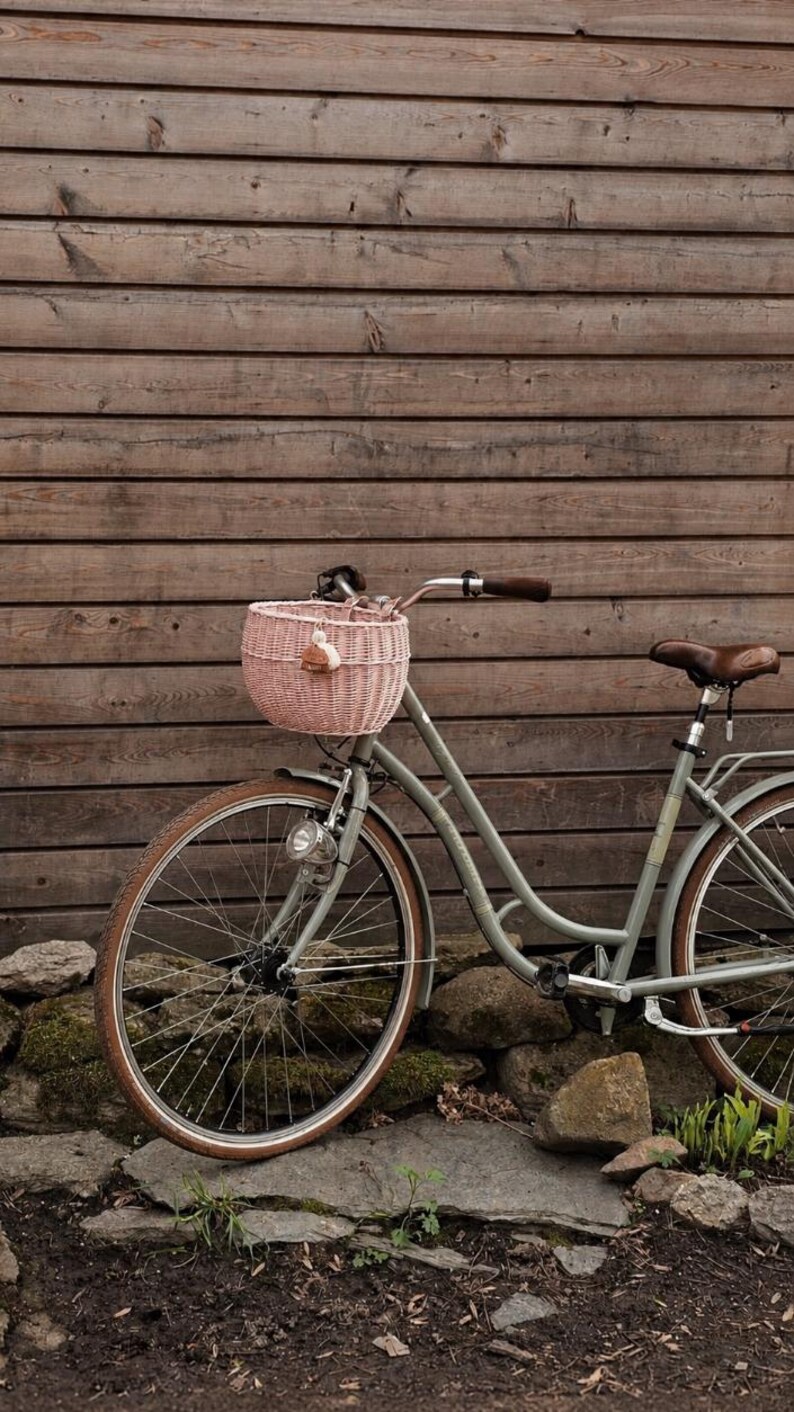 Puede incluir: Una bicicleta de color verde salvia con una cesta de mimbre trenzado de color rosa claro sujeta al manillar. La bicicleta tiene neum&aacute;ticos de goma marr&oacute;n, un sill&iacute;n de cuero marr&oacute;n, manillar de metal plateado con pu&ntilde;os oscuros y un faro.