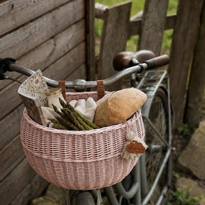 Puede incluir: Una cesta de bicicleta de mimbre tejido de color rosa claro sujeta al manillar de una bicicleta gris. La cesta contiene un manojo de esp&aacute;rragos verdes, una hogaza de pan, un peque&ntilde;o frasco de vidrio y un libro con estampado floral.