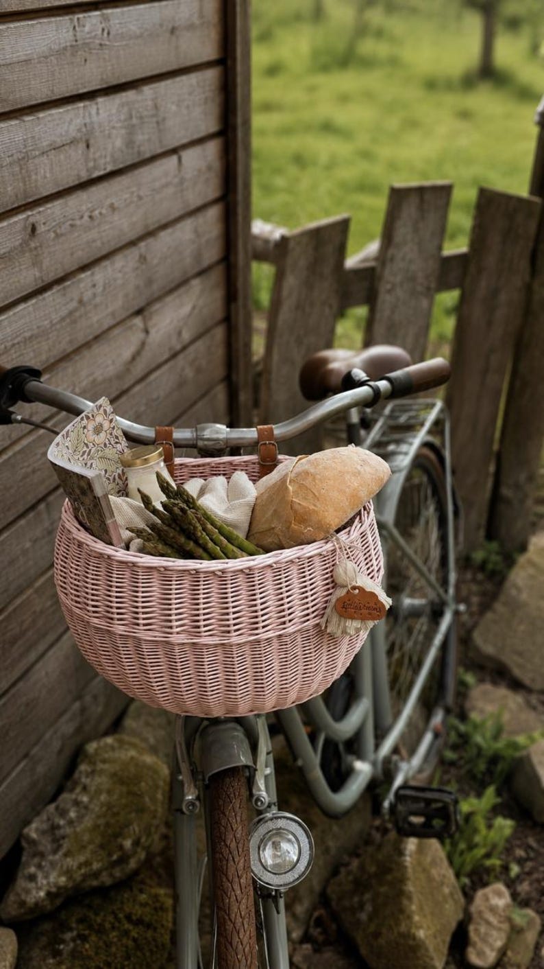 Puede incluir: Una cesta de bicicleta de mimbre tejido de color rosa claro sujeta al manillar de una bicicleta gris. La cesta contiene un manojo de esp&aacute;rragos verdes, una hogaza de pan, un peque&ntilde;o frasco de vidrio y un libro con estampado floral.