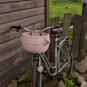 Puede incluir: Una bicicleta de color verde salvia claro con una cesta redonda de mimbre tejido rosa p&aacute;lido sujeta al manillar con correas de cuero marr&oacute;n. La bicicleta incluye un sill&iacute;n marr&oacute;n, un faro delantero plateado y un portaequipajes trasero.