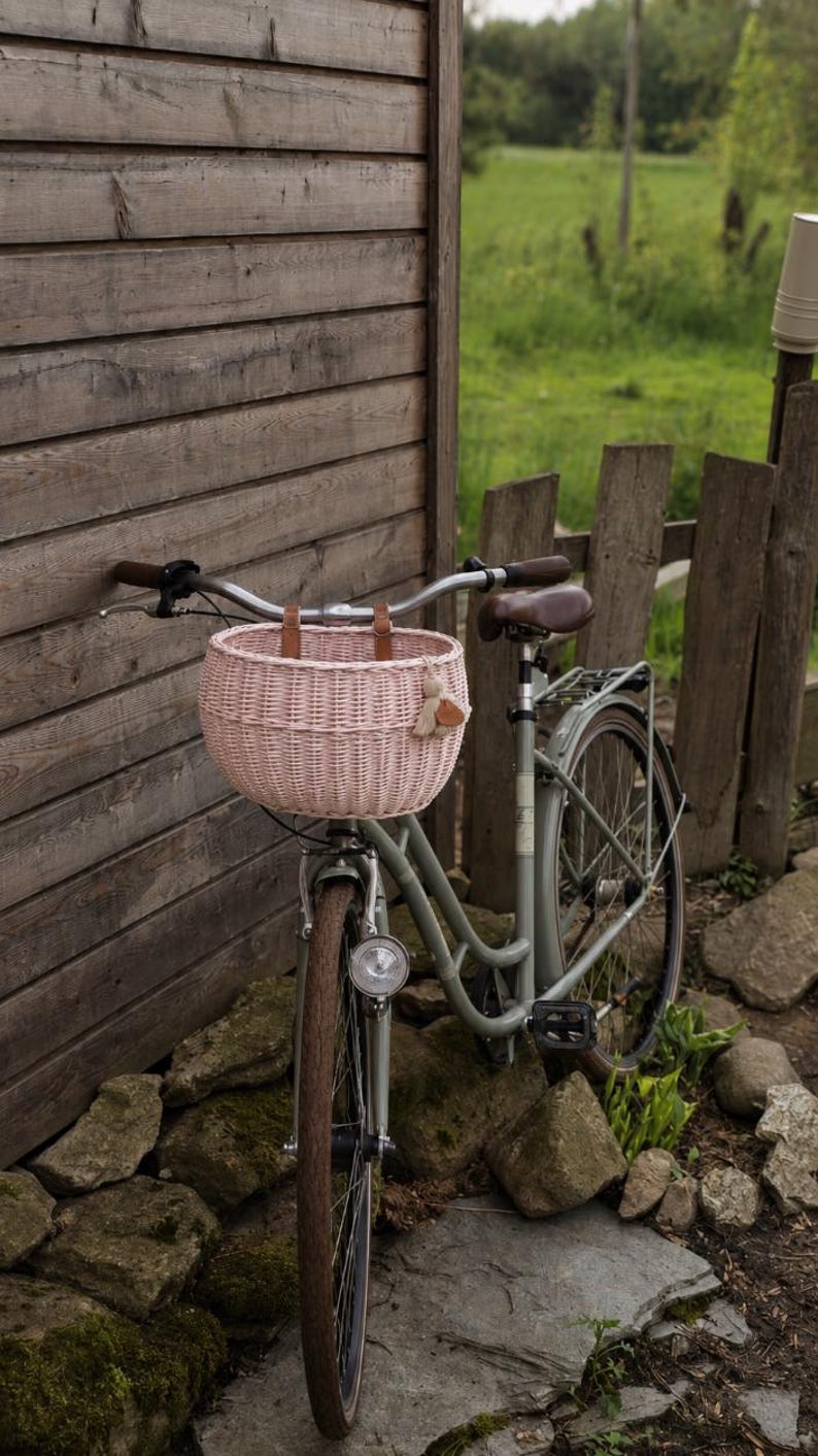 Puede incluir: Una bicicleta de color verde salvia claro con una cesta redonda de mimbre tejido rosa p&aacute;lido sujeta al manillar con correas de cuero marr&oacute;n. La bicicleta incluye un sill&iacute;n marr&oacute;n, un faro delantero plateado y un portaequipajes trasero.