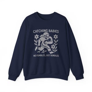 May include: Navy blue sweatshirt with a gray graphic of a football player carrying a baby, surrounded by flowers. The text reads "CATCHING BABIES" above and "NO FUMBLES, JUST BUNDLES" below the graphic.