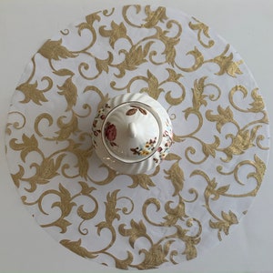 May include: White round tablecloth with a gold floral pattern. A white porcelain sugar bowl with a floral design sits in the center of the tablecloth.