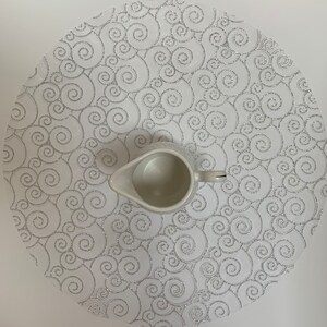May include: White round placemat with a silver swirl pattern. A white ceramic creamer sits on top of the placemat.