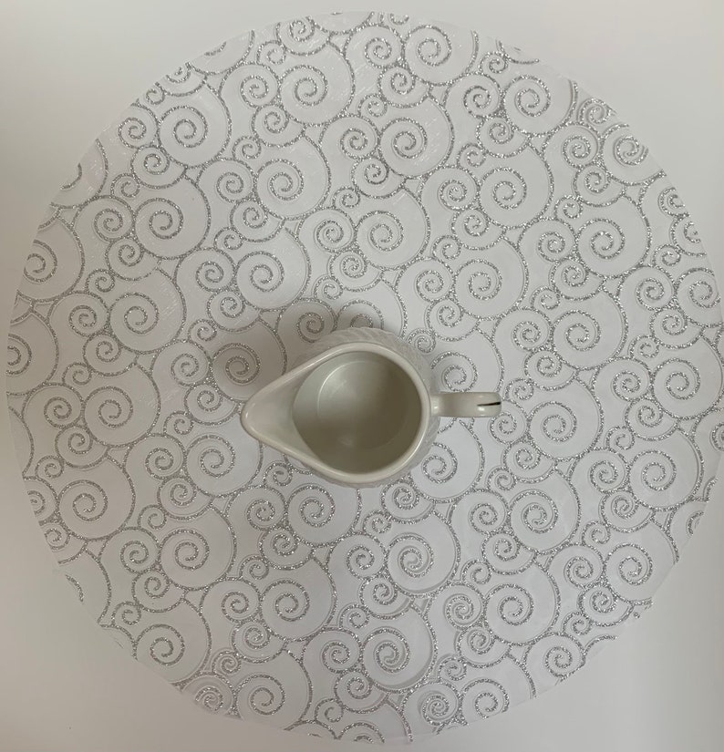 May include: White round placemat with a silver swirl pattern. A white ceramic creamer sits on top of the placemat.