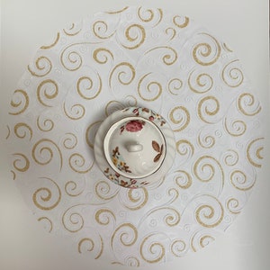 May include: White round table runner with a gold swirl pattern. A white porcelain sugar bowl with floral designs sits in the center of the runner.