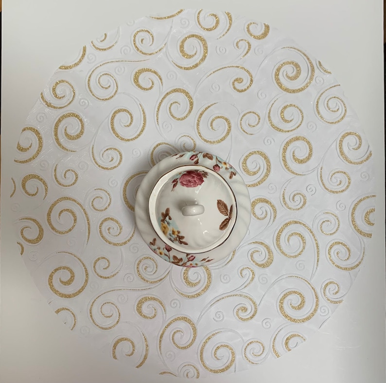 May include: White round table runner with a gold swirl pattern. A white porcelain sugar bowl with floral designs sits in the center of the runner.