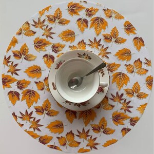 May include: White round placemat with a gold and brown leaf pattern. A white teacup and saucer with a floral design are on top of the placemat.