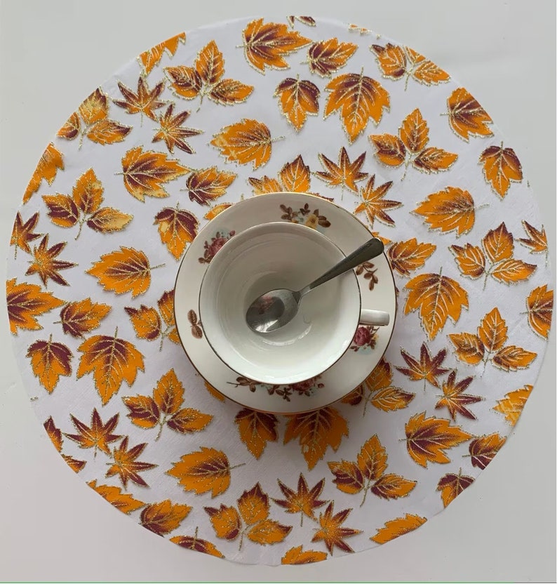 May include: White round placemat with a gold and brown leaf pattern. A white teacup and saucer with a floral design are on top of the placemat.