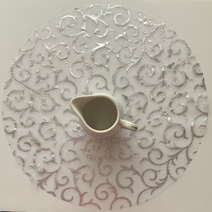May include: A white round table runner with a silver glitter swirl design. A white ceramic creamer sits in the center of the runner.