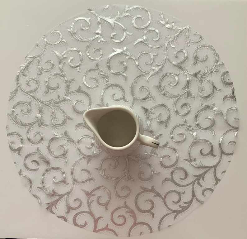 May include: A white round table runner with a silver glitter swirl design. A white ceramic creamer sits in the center of the runner.
